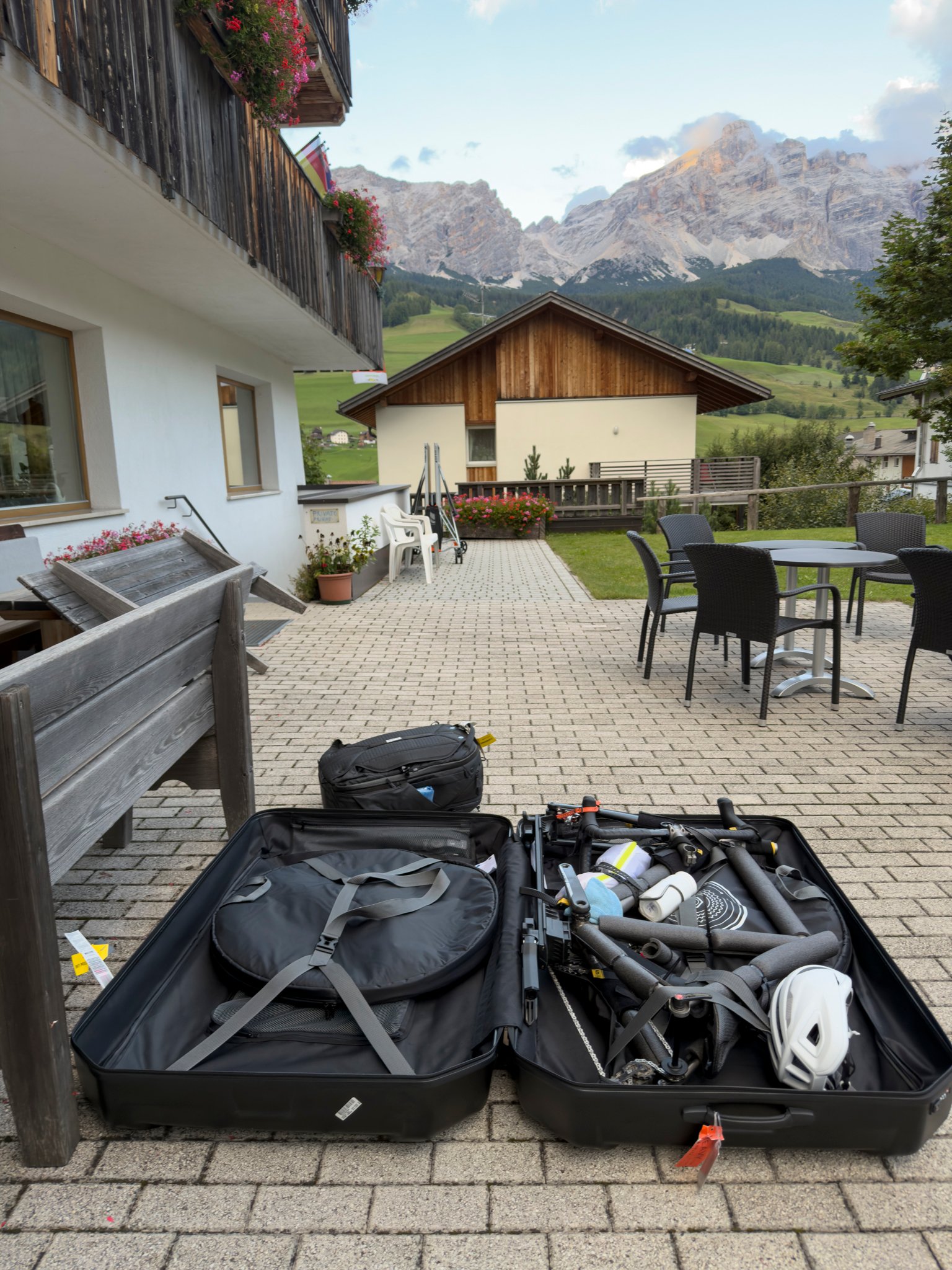 A hard case open on the terrace with the bicycle packed inside, the Dolomites catching last light behind it