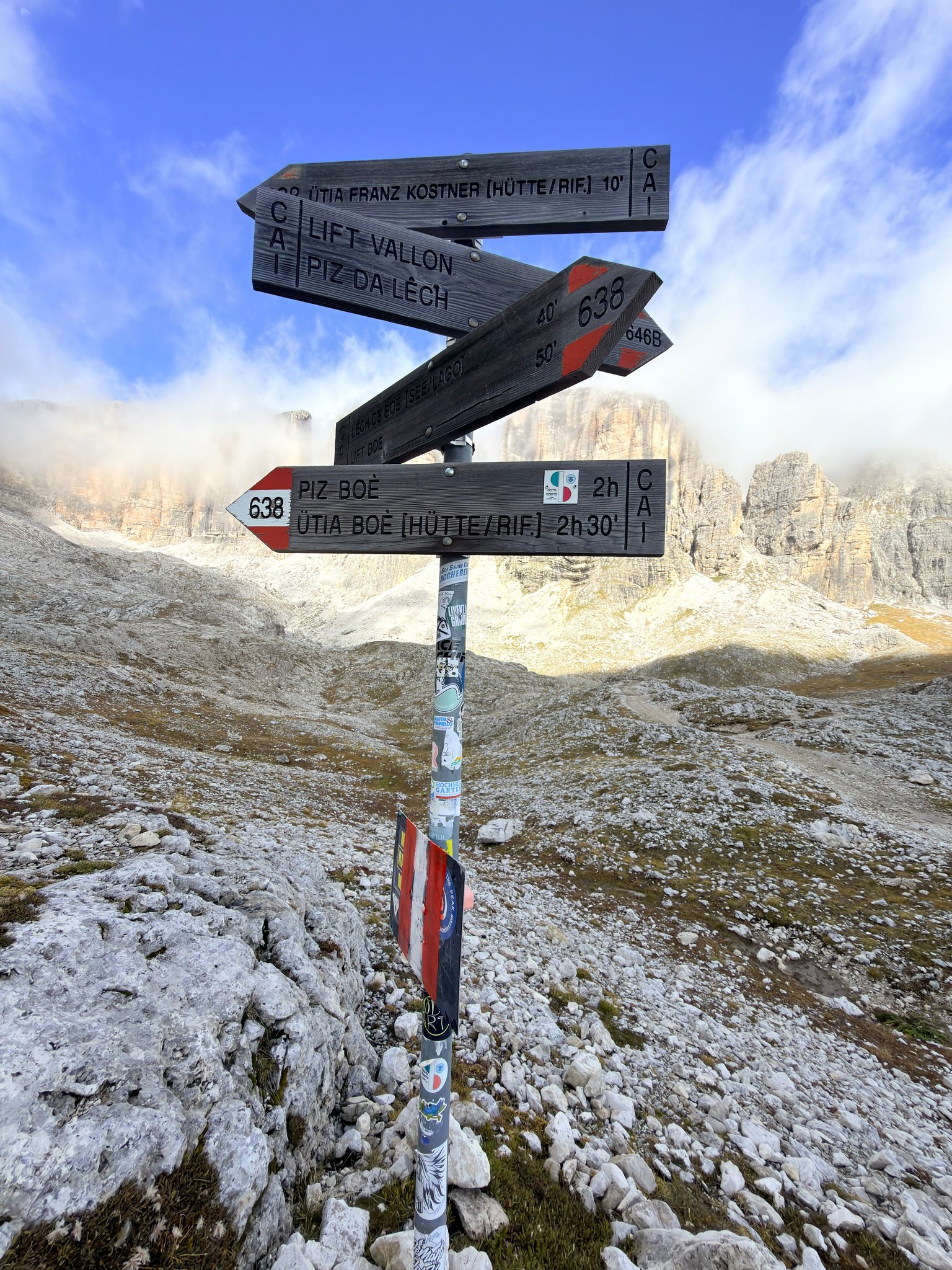 A weathered trail signpost above Rifugio Franz Kostner pointing toward Piz Boè, cloud clearing off the rock